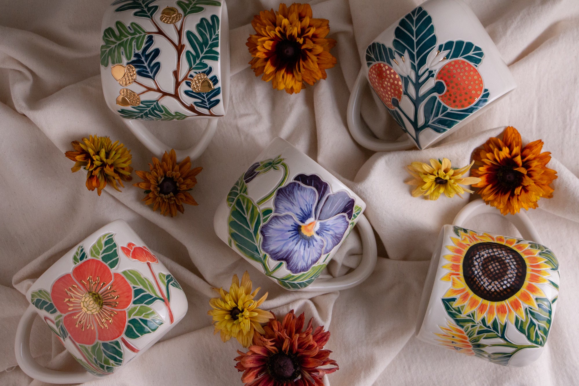Five white ceramic mugs decorated with colorful hand-painted botanical and floral designs, arranged on draped cream fabric with small dried flowers around them.