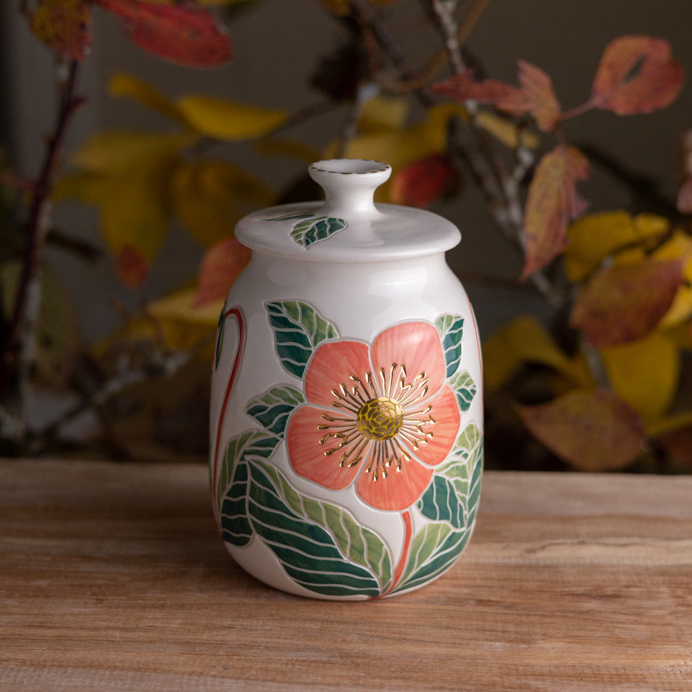 A white ceramic lidded jar with a large pink hellebore flower and gold accents on the front, sitting on a wooden surface with blurred autumn leaves in the background.