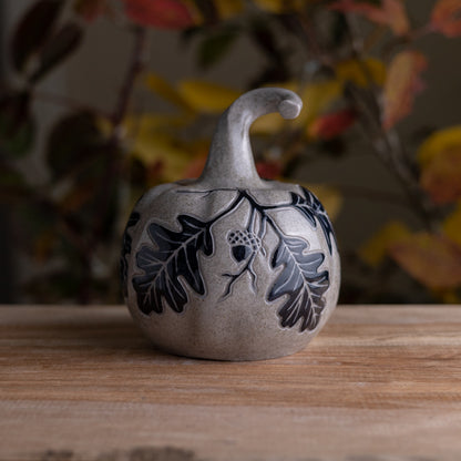 A small decorative ceramic pumpkin figurine with a gray sandstone finish, featuring black hand-painted oak leaves and acorns, displayed on a wooden surface with a blurred autumn background.