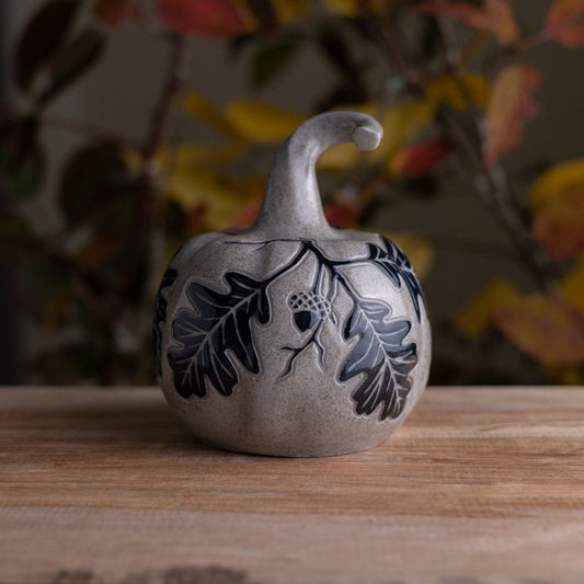 A small decorative ceramic pumpkin figurine with a gray sandstone finish, featuring black hand-painted oak leaves and acorns, displayed on a wooden surface with a blurred autumn background.
